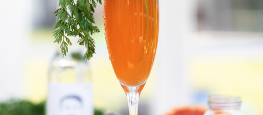 Sektglas mit orangem Cocktail, Möhrenkraut und einer Flasche Schorlefranz Weißweinschorle im Hintergrund